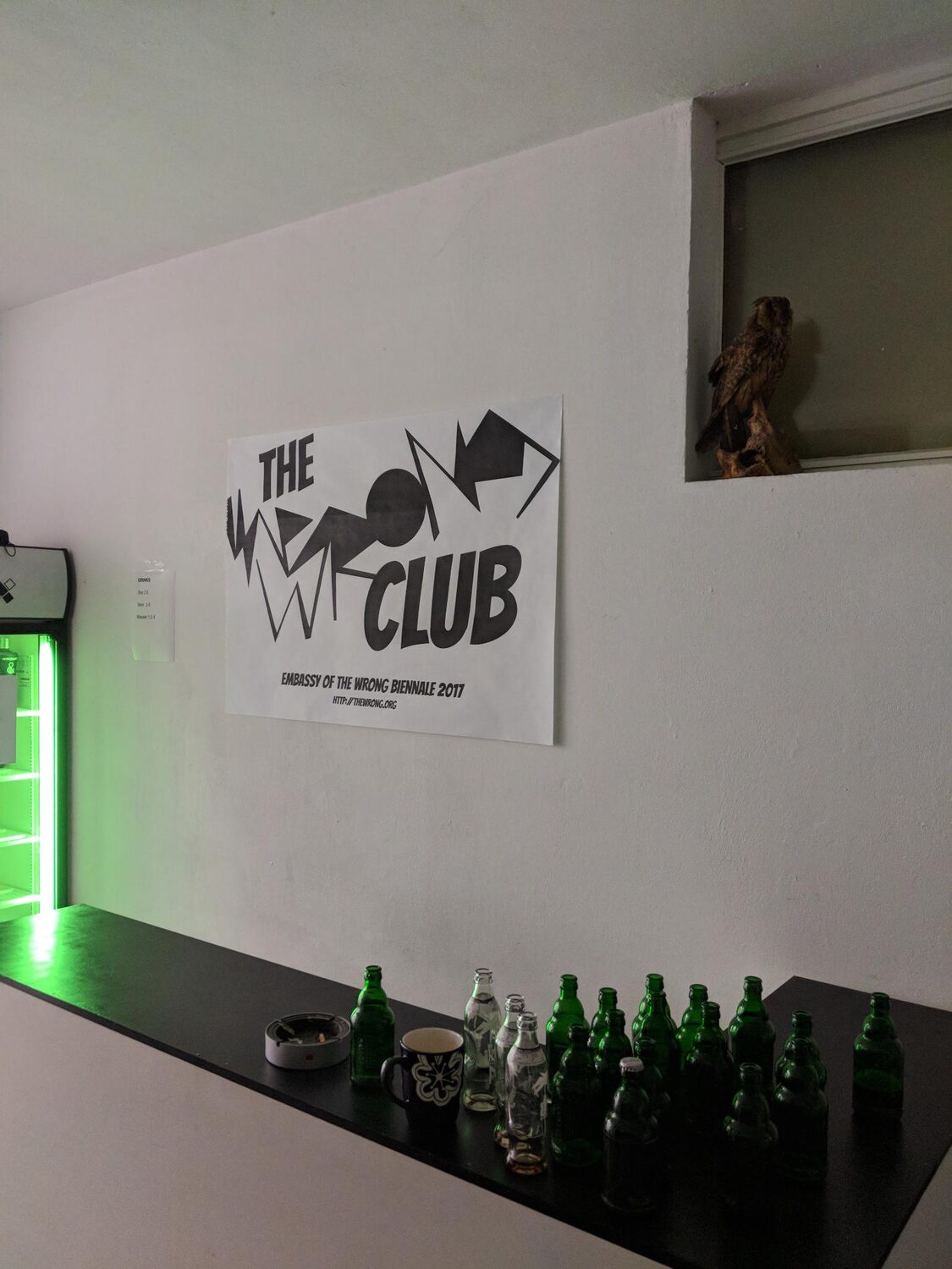 The Wrong Club opening — the club poster 'Embassy of the Wrong Biennale 2017' above the bar, green beer bottles on the counter, a stuffed owl watching from its niche