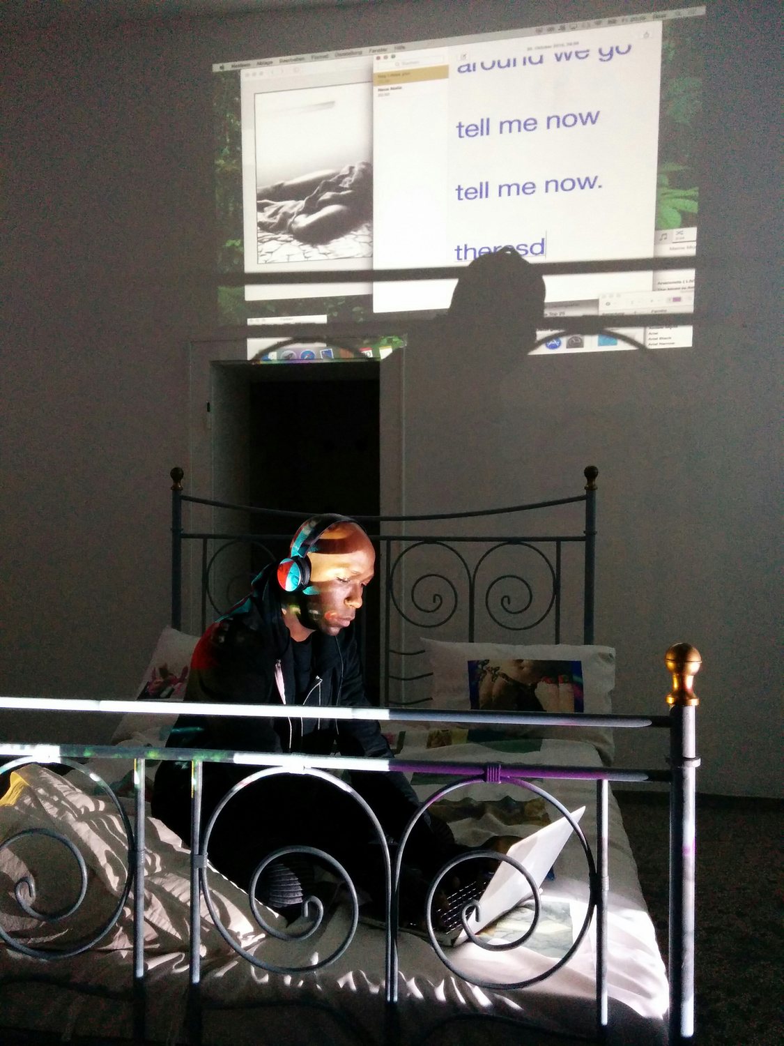 Exploit & Disappear — performer with headphones on the iron bed, 'tell me now' projected
