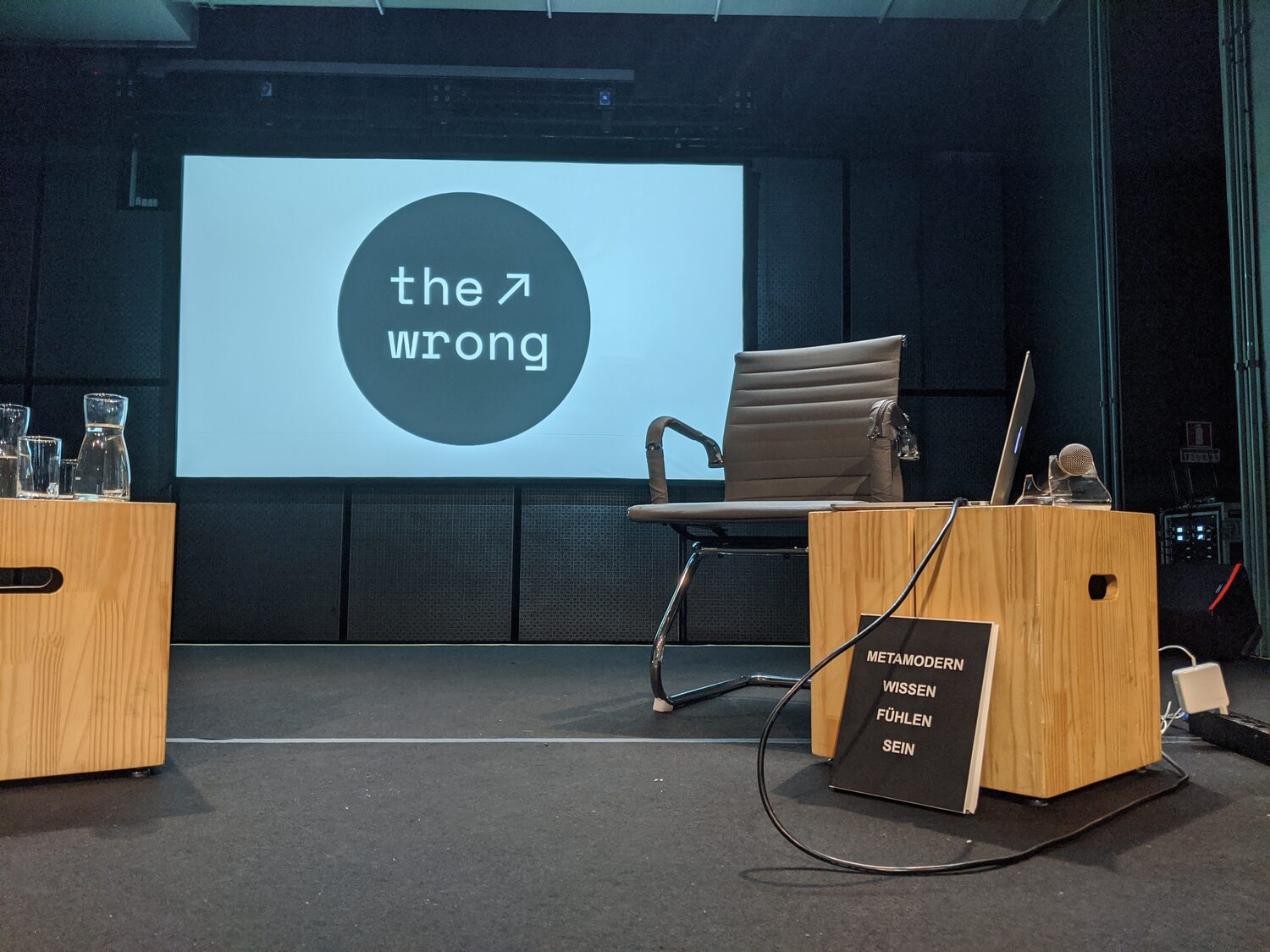 The stage before the lecture at The Wrong Symposium São Paulo — the wrong logo projected on the screen, an empty chair, and the book Metamodern. Wissen Fühlen Sein at the lectern